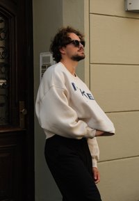 Μπεζ φούτερ με μπλε γράμματα, σε oversized γραμμή, με μαλακή υφή. Φοριέται με μαύρα παντελόνια. Άτομο με σκούρα γυαλιά ηλίου, στέκεται μπροστά σε έναν τοίχο.