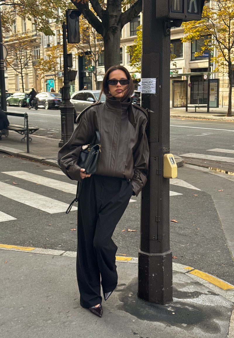 Chaqueta marrón de cuero con cuello alto, mangas oversized y bolsillo; combinada con pantalones negros sueltos y tacones negros puntiagudos. Bolso negro en la mano.