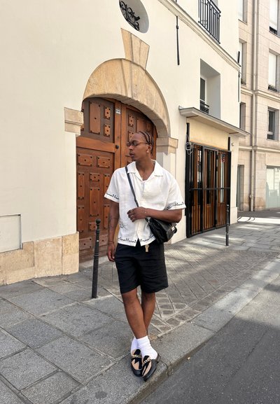 Camisa blanca de manga corta con ribete negro, pantalones cortos negros y zapatos negros con un acento de cadena, acompañados de gafas de sol y un pequeño bolso negro.
