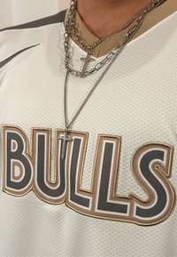 Biała koszulka sportowa z teksturowanej tkaniny, z dużym napisem "BULLS" w szaro-złotym kolorze. Srebrne łańcuchy z krzyżem wiszącym na wierzchu.