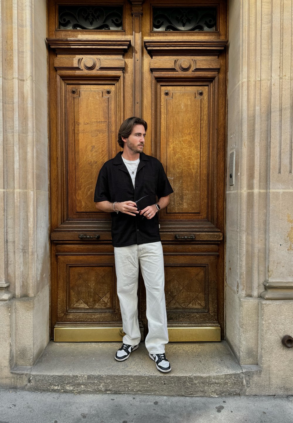 Μαύρο κοντομάνικο πουκάμισο, λευκό T-shirt, ανοιχτόχρωμο παντελόνι και μαύρα με λευκά αθλητικά παπούτσια. Στημένος μπροστά από περίτερες ξύλινες πόρτες.