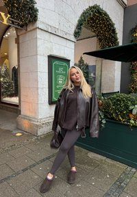 Brązowa, skórzana, oversize'owa kurtka, ciemnobrązowy, puszysty sweter, szare legginsy oraz brązowe buty wsuwane z białym przeszyciem, stojące obok świątecznej zieleni.