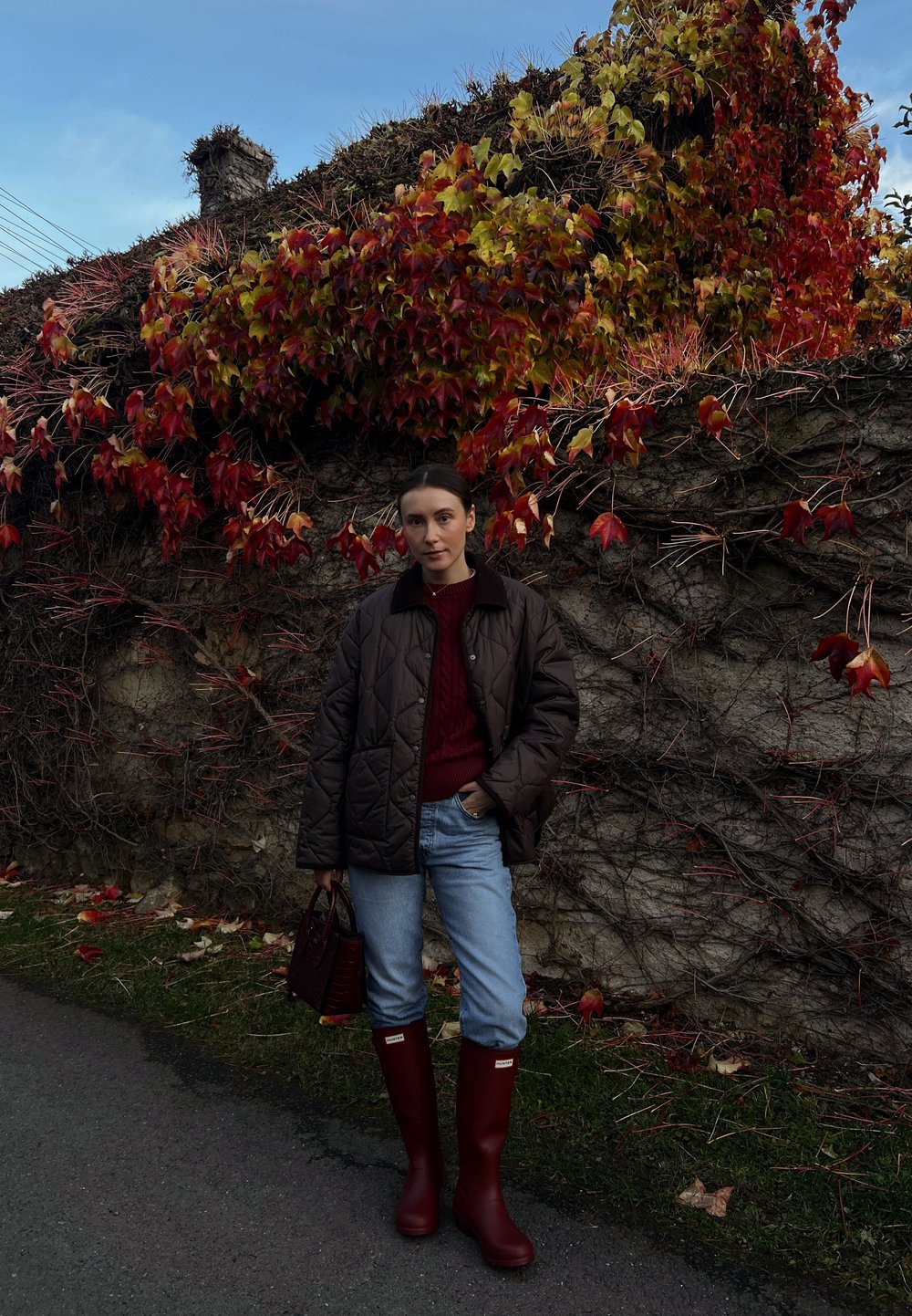 Hnedá prešívaná bunda, bordový sveter, modré rifle a červené gumáky. Drží tmavú kabelku s textúrovaným vzorom. Na pozadí jesenné lístie.