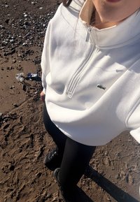 Personne portant un sweat à capuche blanc zippé et un legging noir debout sur un sol sablonneux et rocheux, avec des déchets éparpillés à proximité.