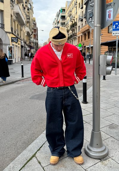 Chaqueta corta roja con rayas blancas, pantalones de mezclilla oscuros y holgados, zuecos beige, y una diadema marrón y negra. Entorno urbano con edificios.