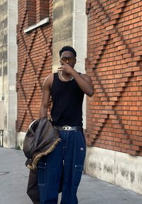 Camisola preta sem mangas, jeans de perna larga azul com bolsos, casaco de couro escuro com acabamento em pele, e fundo único de parede de tijolos castanha.