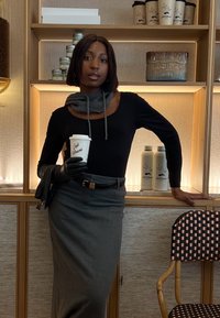 Maglietta nera a maniche lunghe, sciarpa grigia, gonna a tubino grigia e guanti di pelle neri. Tiene una tazza di caffè davanti a un'esposizione in legno con oggetti.