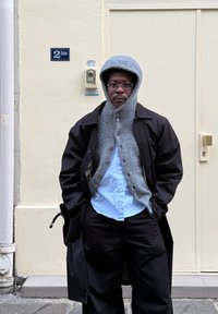 Pelēks adīts džemperis ar kapuci, kārtots zem melna mēteļa, gaiši zila krekla un tumšu biksu. Stāv pret gaiši brūnu sienu ar durvīm.