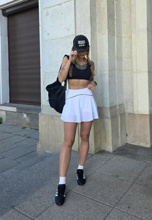 Gorra negra con letras blancas, top corto negro, falda corta blanca de patinadora con una línea de acento, zapatillas negras y una mochila negra.