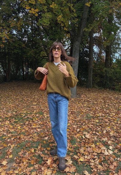 Suéter de punto verde oliva, camisa clara por debajo, jeans de tiro alto azules, botas de tobillo marrones, sosteniendo un bolso de mano tejido naranja. Hojas de otoño alrededor.