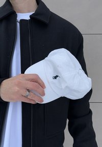 Personne portant une veste noire et une chemise blanche, tenant une casquette blanche avec un petit logo noir, devant un mur gris carrelé.