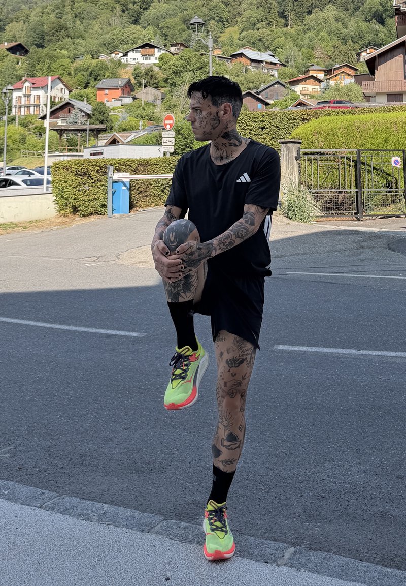 Mężczyzna z tatuażami, ubrany w czarny strój sportowy i neonowe buty do biegania, rozciąga nogę na ulicy w górskiej wiosce z domami i zielenią w tle.
