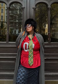 Červená košeľa s grafickou potlačou, zelená károvaná kravata, vrstvené so sivým kabátom. Doplnky zahŕňajú veľké čierne okuliare a čiernu kožušinovú čiapku.