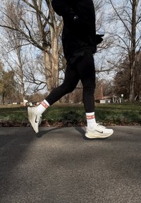 Bijele Nike tenisice za trčanje s debelim potplatom, uparene s crnim tajicama i bijelim čarapama s crvenim i bijelim detaljima. Osoba u pokretu na pločniku.