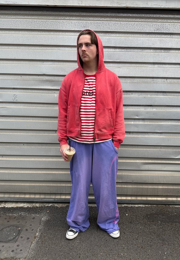 Young man in a red hoodie and purple pants stands against a gray metal wall holding a drink with a straw, looking to the side.