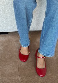 Crvene lakirane Mary Jane cipele s remenčićem preko gornjeg dijela stopala, nošene uz svijetloplave traperice s manžetama. U pozadini mekani bež tepih.