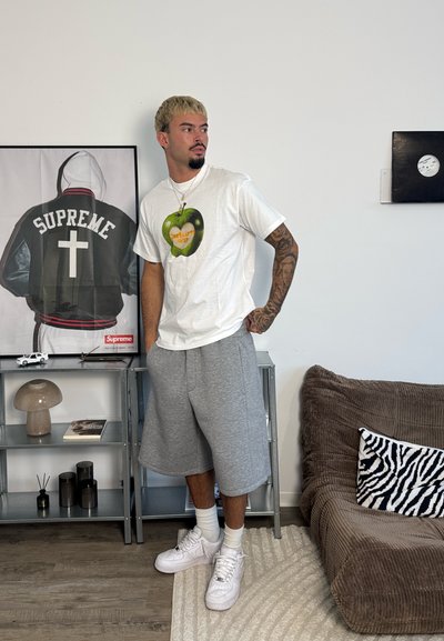 Hombre con el brazo tatuado que lleva camiseta blanca y pantalones cortos grises, de pie junto a estanterías metálicas con foto enmarcada de una chaqueta Supreme y objetos decorativos.