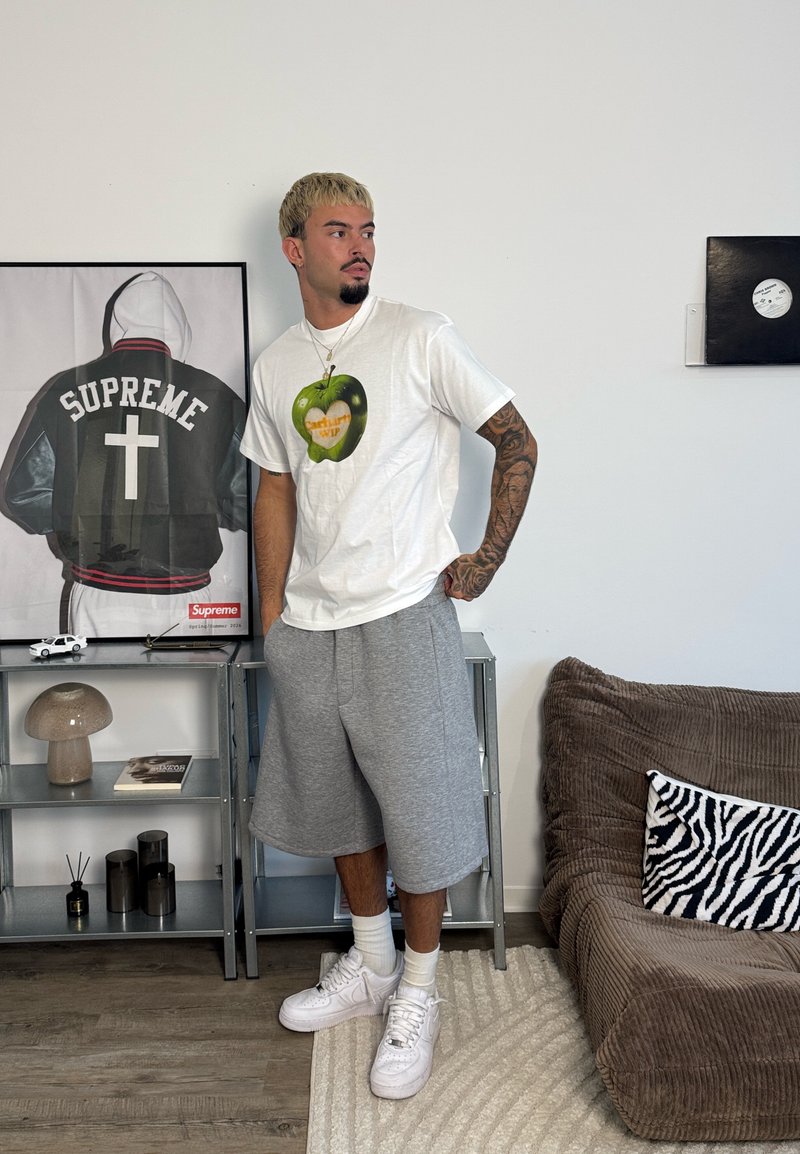 Homme avec un bras tatoué portant un t-shirt blanc et un short gris se tient à côté d'étagères métalliques avec une photo encadrée d'une veste Supreme et des objets décoratifs.