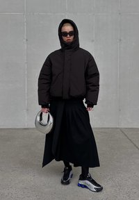 Casaco acolchoado preto com capuz, saia preta plissada e ténis pretos com detalhes azuis. A segurar um capacete prateado refletor.