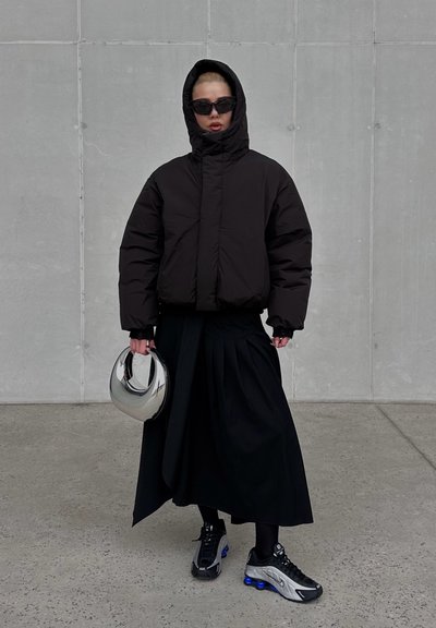 Chaqueta acolchada negra con capucha, falda negra plisada y zapatillas negras con acentos azules. Sostenido un casco plateado reflectante.