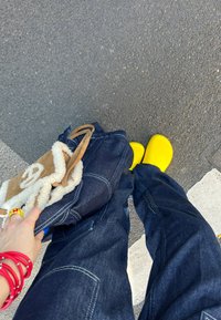 Rumene klompe, modri jeansi s vidnimi šivi in svetlo rjava torba s ovitkom iz ovčje kože, ki jo drži roka z rdečimi zapestnicami.
