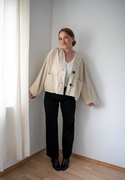 Chaqueta de lino beige recortada con dos bolsillos frontales, botones negros, llevada sobre una blusa blanca y combinada con pantalones negros y botas de tacón.
