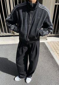 Casaco preto com fecho zip e gola alta, com três riscas brancas nos ombros, combinado com calças largas pretas e ténis brancos.