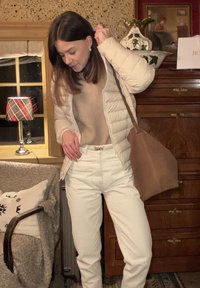 Doudoune beige clair, pull en maille couleur fauve, pantalon blanc taille haute et sac marron. En arrière-plan, une lampe à carreaux et du mobilier en bois.