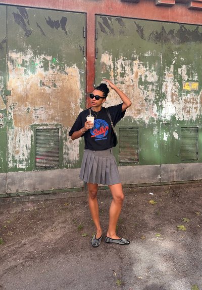 Camiseta gráfica negra con estampado azul y rojo, mini falda plisada gris, zapatos negros tipo slip-on adornados con tachuelas blancas. Vaso de plástico en la mano.