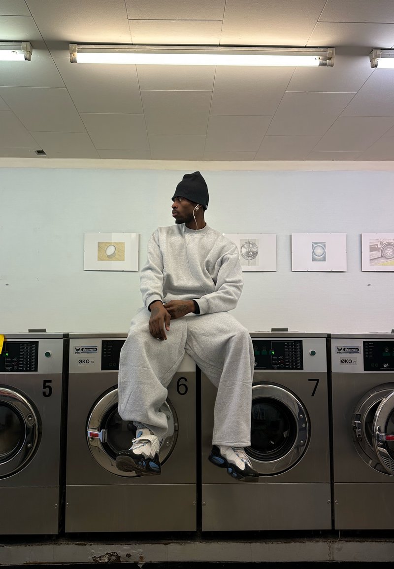 Homme portant un pantalon de survêtement gris, un sweat-shirt, un bonnet noir, et des baskets, assis sur une machine à laver industrielle dans une laverie automatique, regardant sur le côté.