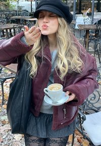 Bordeaux farvet faux læderjakke med blødt foer, grå sweater, plisseret stribet nederdel, holder en hvid kaffekop på underkop, blæser et kys.