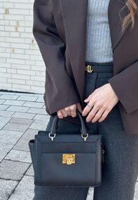 Γυναίκα κρατώντας μαύρη δερμάτινη τσάντα με χρυσό κούμπωμα, φοράει καφέ σακάκι, γκρι ριμπ τοπ, γκρι παντελόνια και χρυσή αγκράφα ζώνης με αρχικά «RL».