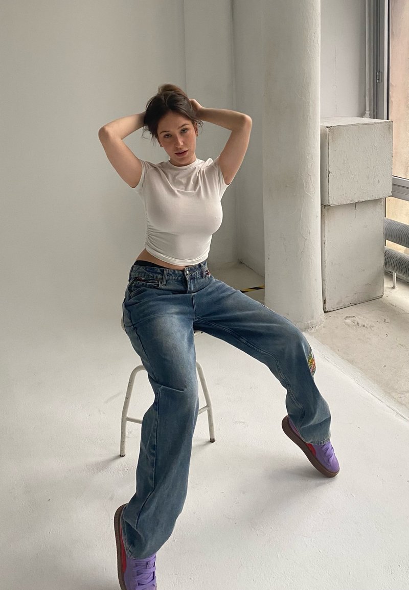 T-shirt ajustée blanche, jeans bleu clair surdimensionnés avec détails en patch, et baskets violettes. Modèle assis sur un tabouret blanc contre un fond neutre.