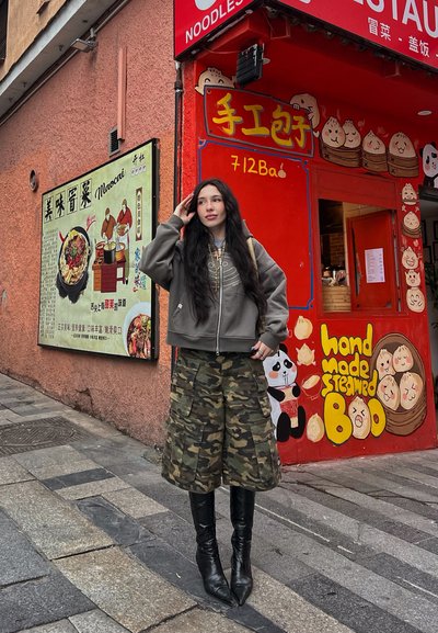 Sudadera gris con cremallera, falda cargo de camuflaje y botas negras hasta la rodilla. Fondo de restaurante colorido con ilustraciones de comida en estilo de dibujos animados.