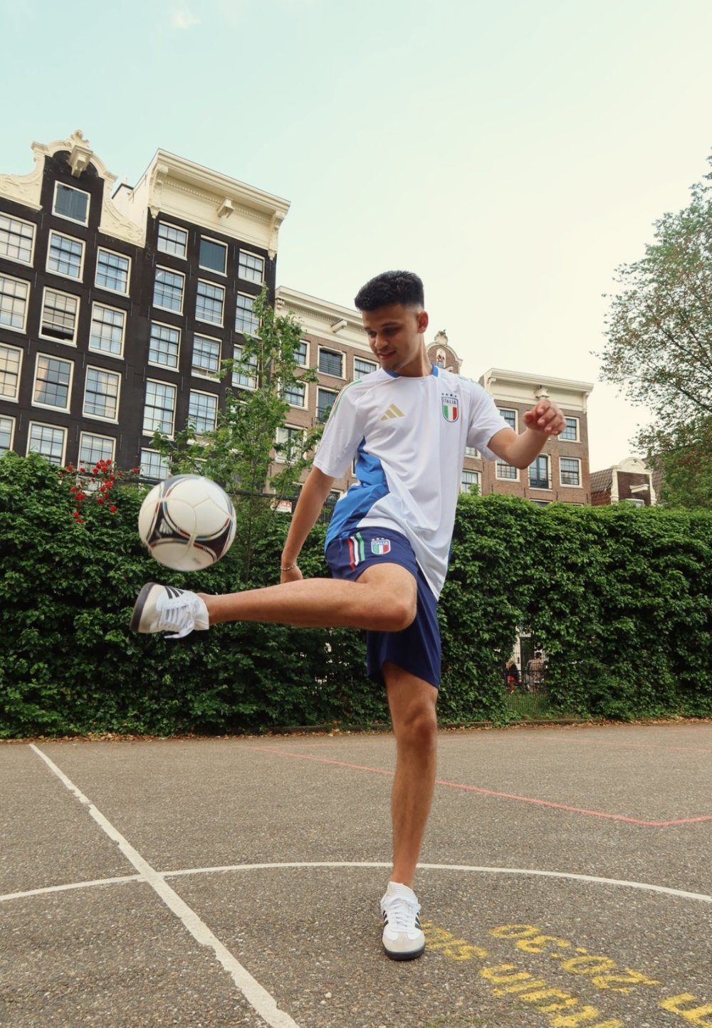 Bílé a modré sportovní tričko s italským emblémem, modré šortky, žonglující s černobílým fotbalovým míčem na dlážděném hřišti obklopeném zelení.