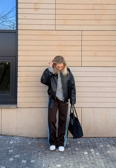 Mujer con cabello rubio que lleva chaqueta de cuero negra, camisa a rayas, pantalones negros con rayas azules, zapatillas blancas, sosteniendo un bolso tote negro, de pie junto a una pared beige.