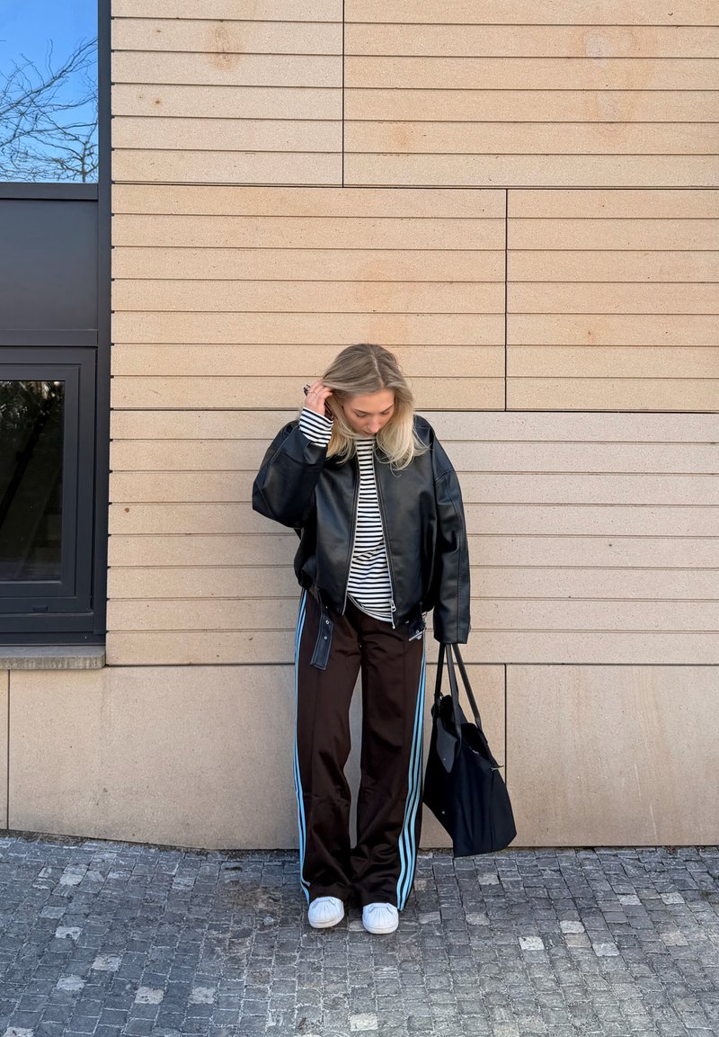 Femme aux cheveux blonds portant une veste en cuir noire, une chemise à rayures, un pantalon noir avec des rayures bleues, des baskets blanches, tenant un sac fourre-tout noir, debout près d'un mur beige.