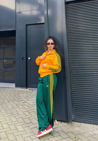 Chándal atlético en vibrante amarillo y verde, con tres rayas, combinado con zapatillas rojas. Gafas de sol elegantes y una pose casual.