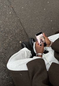 Des chaussures noires à semelles épaisses, un pantalon de survêtement gris et une veste marron surdimensionnée. Une main tenant un smartphone prend un selfie sur un trottoir texturé.