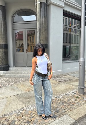 Top sin mangas blanco, jeans de pierna ancha de talle alto en azul claro, tacones negros con estampado y un bolso estampado, de pie en una calle de adoquines.