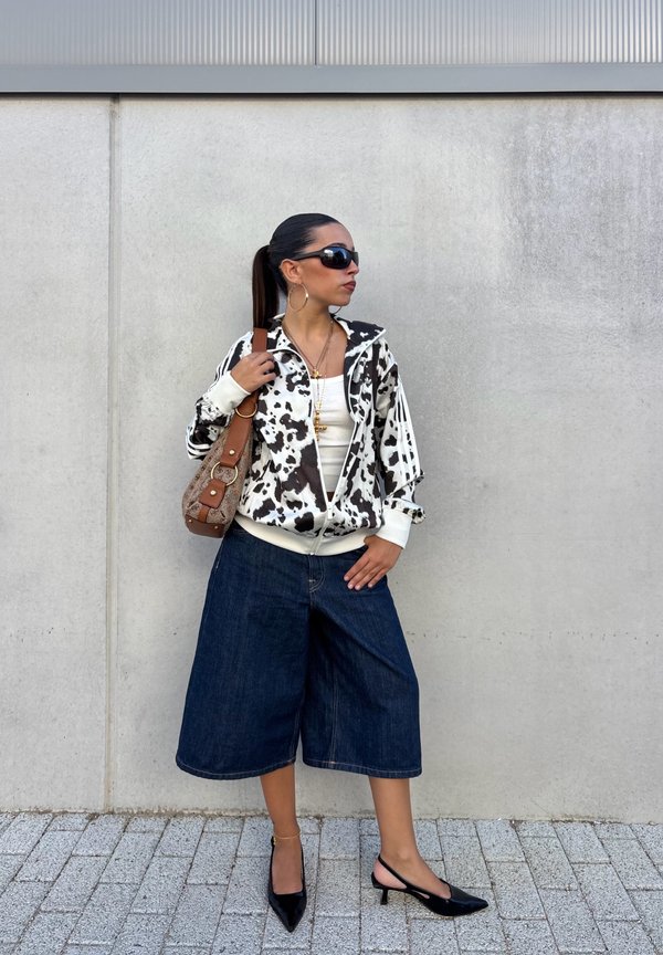 Femme portant une veste à imprimé vache noir et blanc, des culottes en denim, des talons noirs, de grandes boucles d’oreilles, des lunettes de soleil, et tenant un sac à main marron.