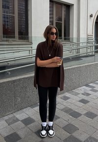 Maglione marrone a coste oversize con collo alto, leggings neri e sneakers bianco e nere. Tiene una tazza di caffè, in piedi su pavimentazione in piastrelle.