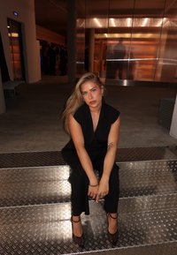 Svart jumpsuit med korta ärmar, sittande på metalliska steg med diamantmönster, bärande svarta remsandaler, med långt blont hår och minimala accessoarer.