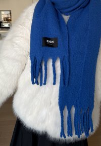 Kék kötött sál rojtos végekkel, puha anyagból készült, fehér műszőrme kabát előtt bemutatva. A címkén az "ZIGN STUDIO" felirat olvasható.
