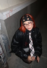 Jeune personne aux cheveux roux portant une veste en cuir noir, un bandeau à trous ronds, une cravate à carreaux et des lunettes, assise sur un trottoir la nuit.