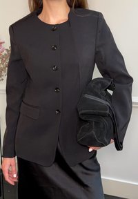 Γυναίκα που φοράει μαύρο σακάκι με κουμπιά και σατέν φούστα, κρατώντας μια μαύρη τσάντα από σουέντ, πάνω σε απλό φωτεινό φόντο.