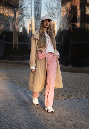 Gabardina beige, suéter de punto blanco, pantalones rosas, zapatillas blancas y un bolso cruzado rosa. Cabello largo y liso y una gorra clara.