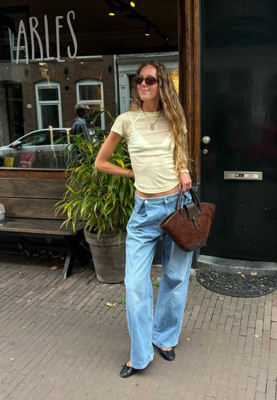 Top de manga corta, color amarillo claro, combinado con vaqueros anchos azules. Sosteniendo un bolso de ante marrón con detalles negros. Planta verde al fondo.