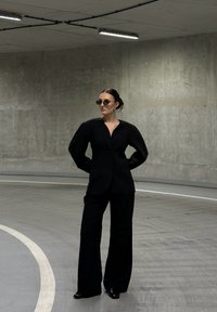 Žena v černém oversized blejzru a širokých kalhotách stojí v zakřivené betonové parkovací garáži, má na sobě malé černé sluneční brýle a černé boty.