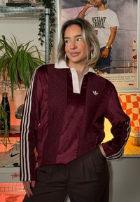 Mørkerød velour langermet polo med hvit krave og tre hvite striper på ermene. Inkluderer teksturerte detaljer i stoffet.